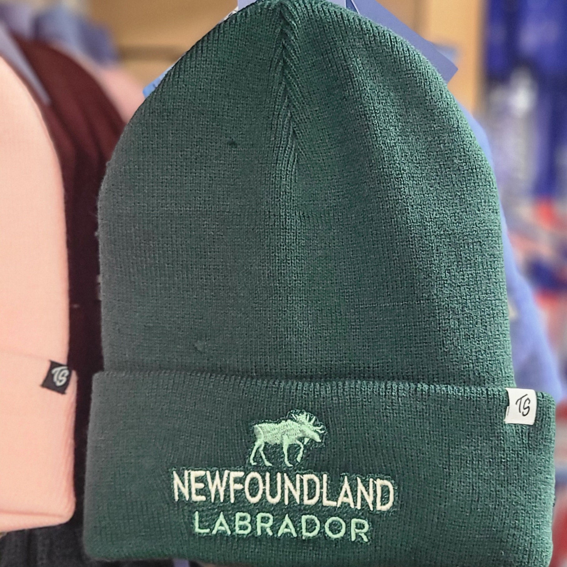 Green beanie with 'Newfoundland Labrador' text held by a person in a clothing store.
