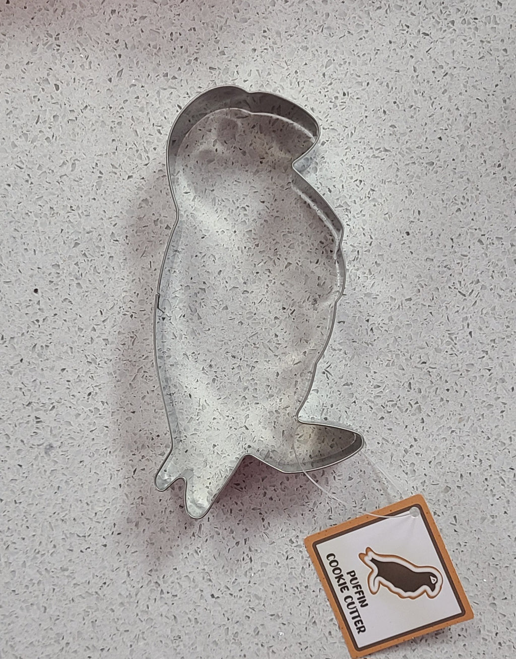 Metallic penguin-shaped cookie cutter on a gray surface with a brand label in the corner.