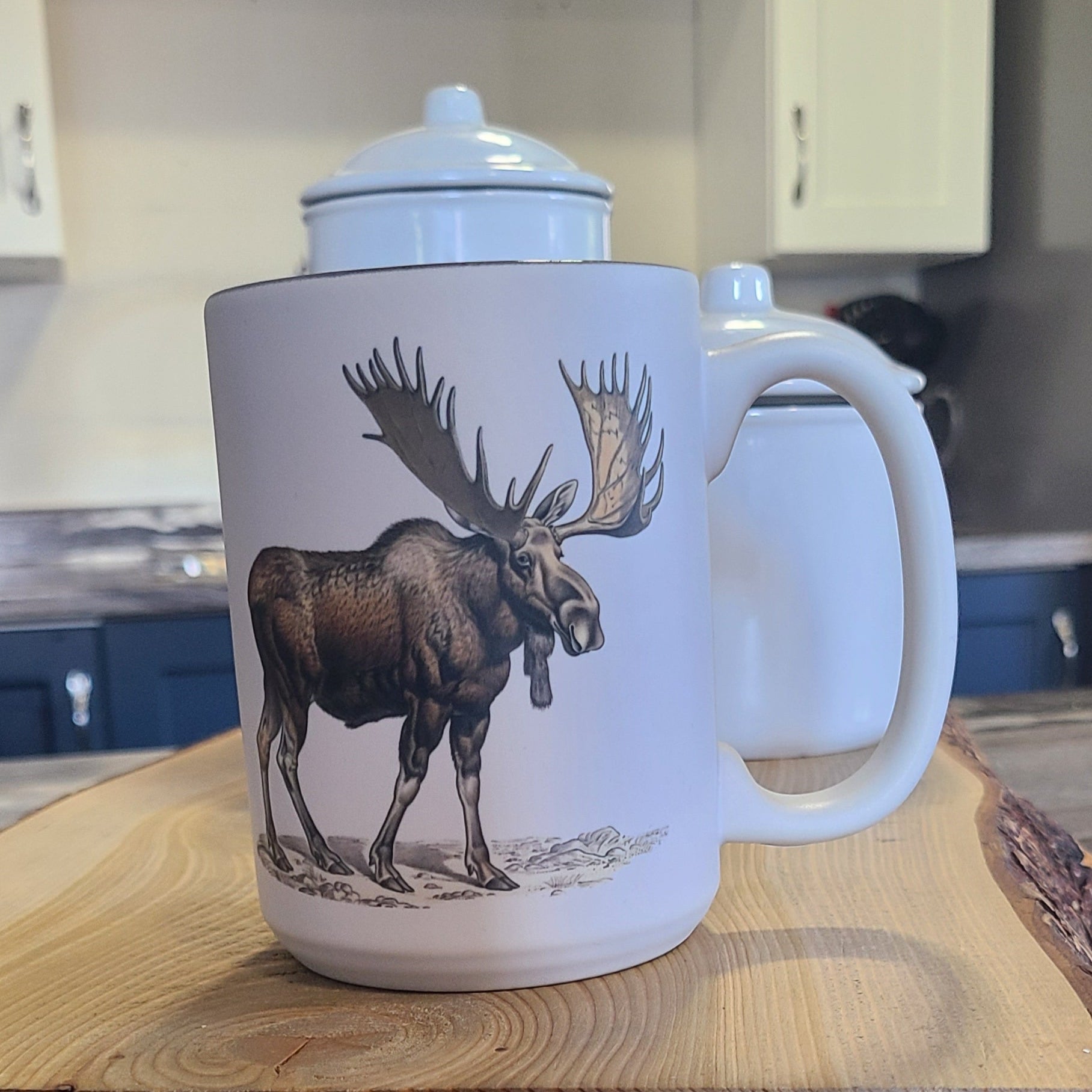 Ceramic mug with detailed illustration of a moose standing on a rocky ground, Newfoundland style, on a kitchen counter.