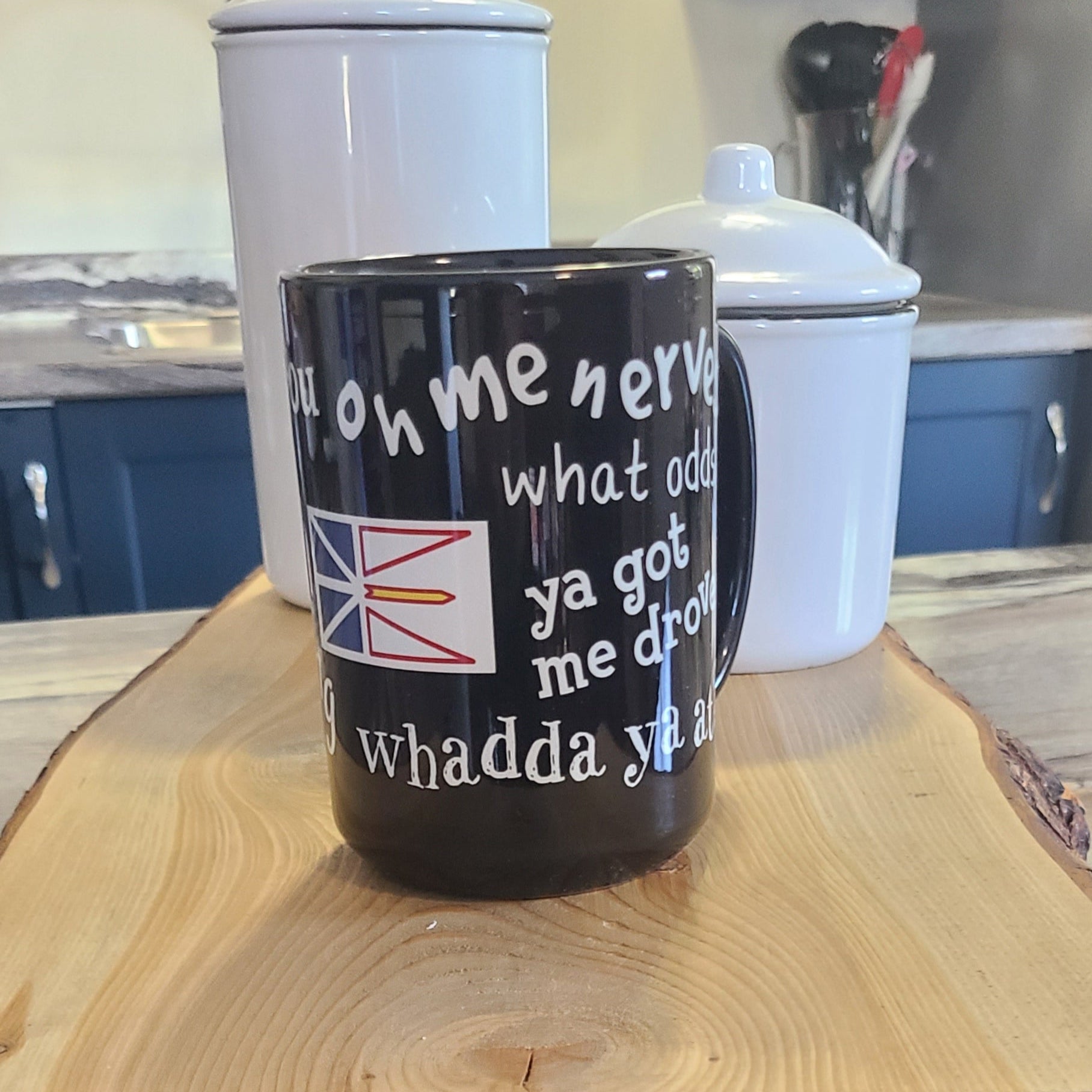 Black ceramic mug with Newfoundland sayings like “What odds” and “Ya got me drove,” alongside the Newfoundland flag, sitting on a wooden board.