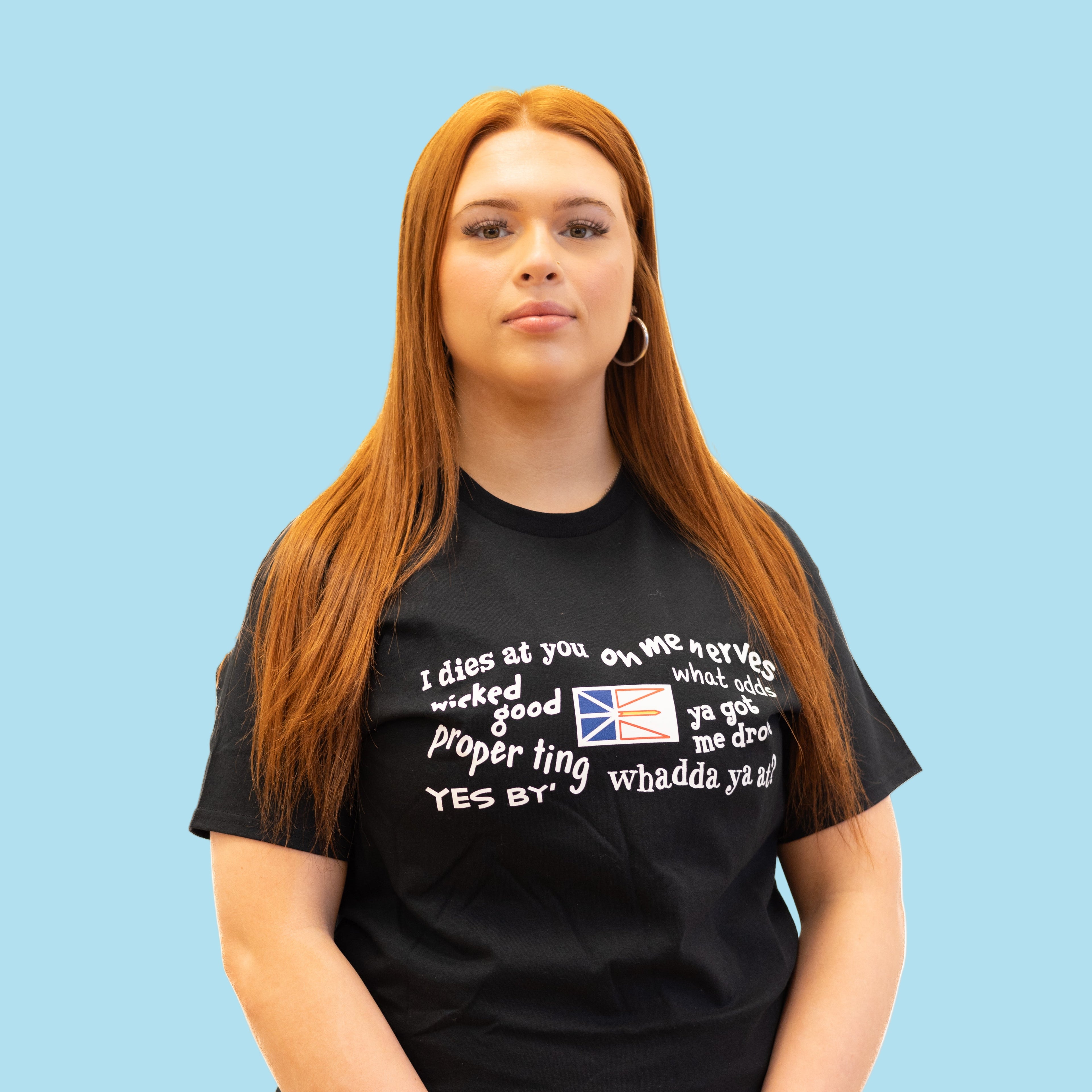 Person wearing a black Newfoundland Sayings T-shirt with fun local phrases and the Newfoundland flag printed on the front.