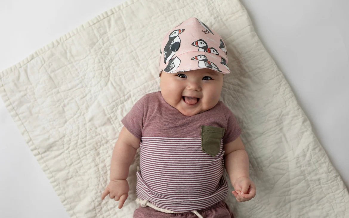Baby wearing a pink baseball cap with Newfoundland's puffin pattern on a beige blanket.