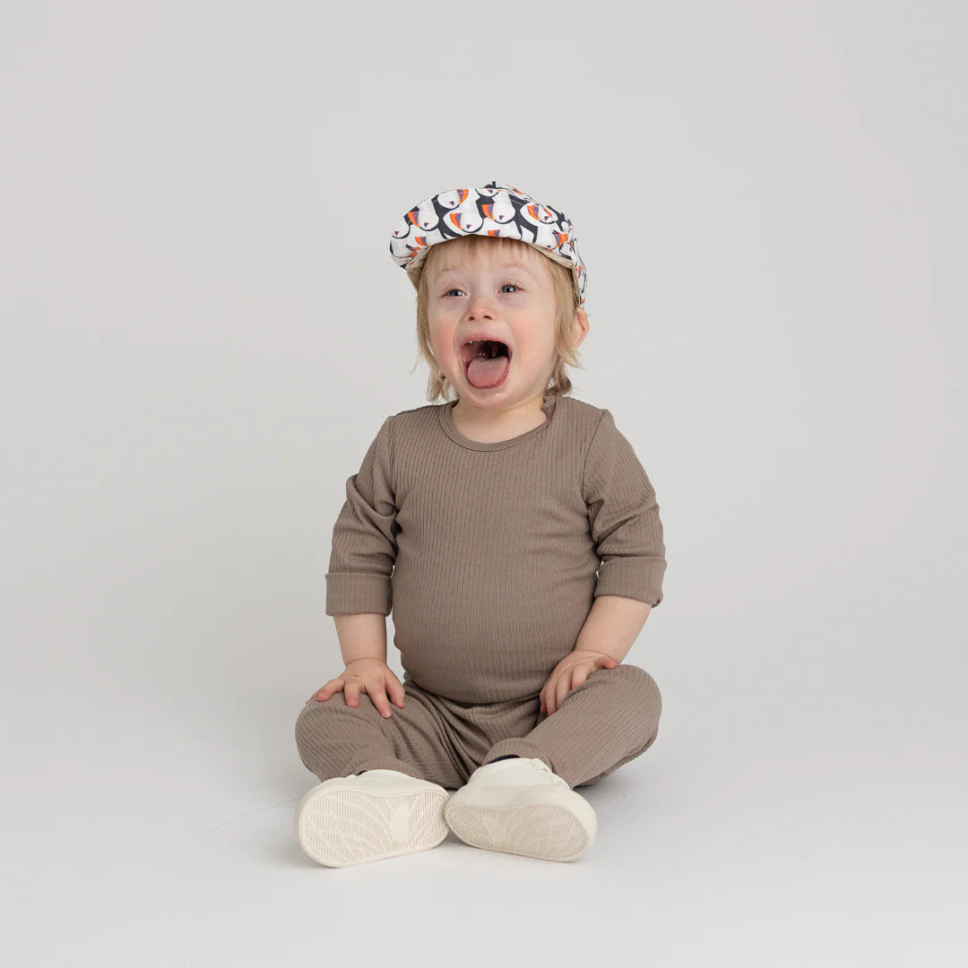 Toddler wearing a brown outfit and Puffin Baseball Cap with white base hat on a plain background.
