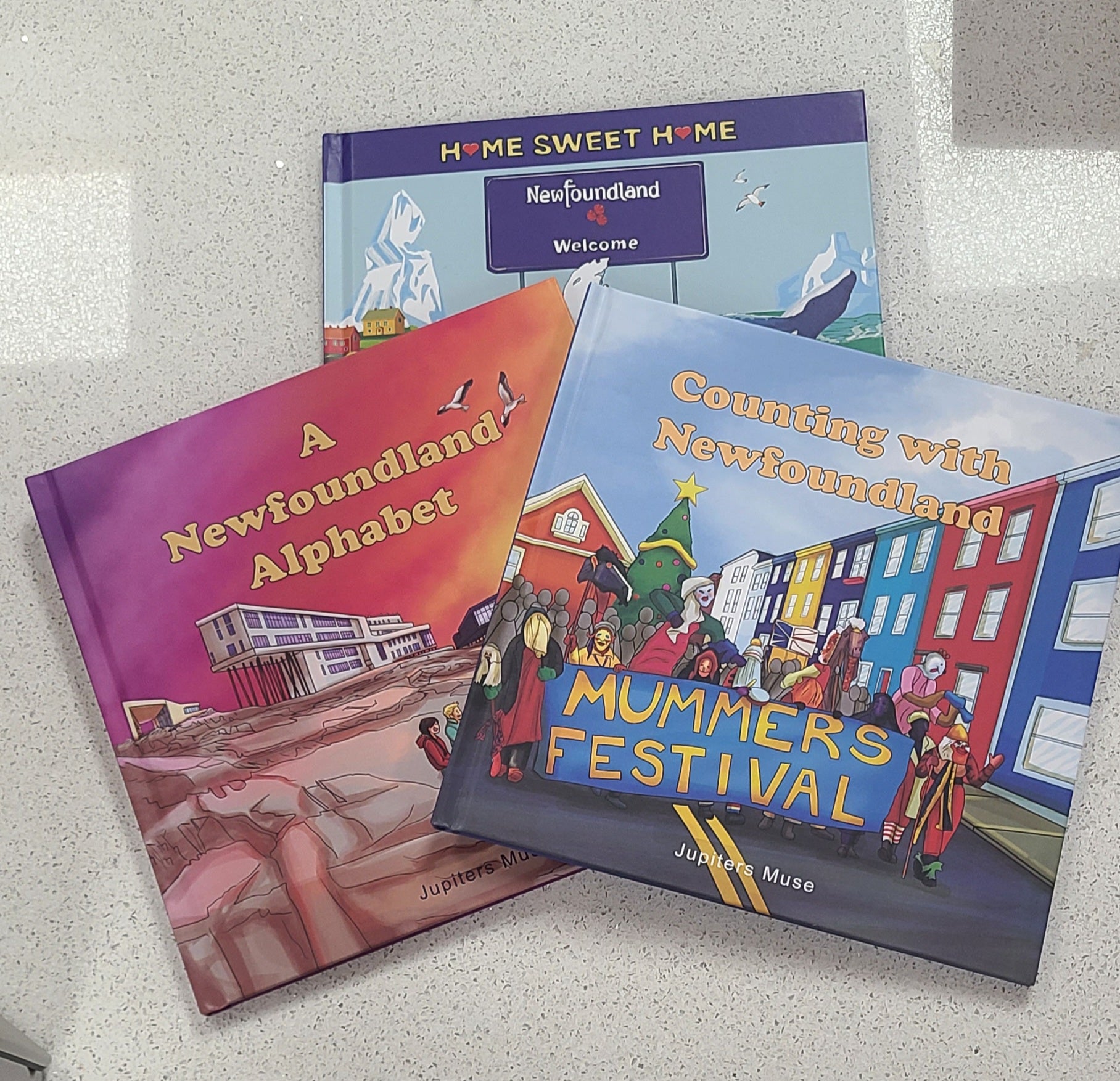 Three books titled 'A Newfoundland Alphabet', 'Home Sweet Home Newfoundland Welcome', and 'Counting with Newfoundland Mummer's Festival' on a textured surface.